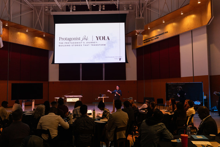 A presenter addresses a seated audience beneath a Protagonist Ink and YOLA title slide.