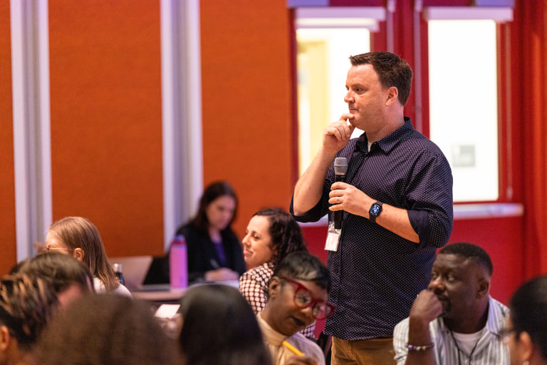 A man holding a microphone listens among seated workshop participants.