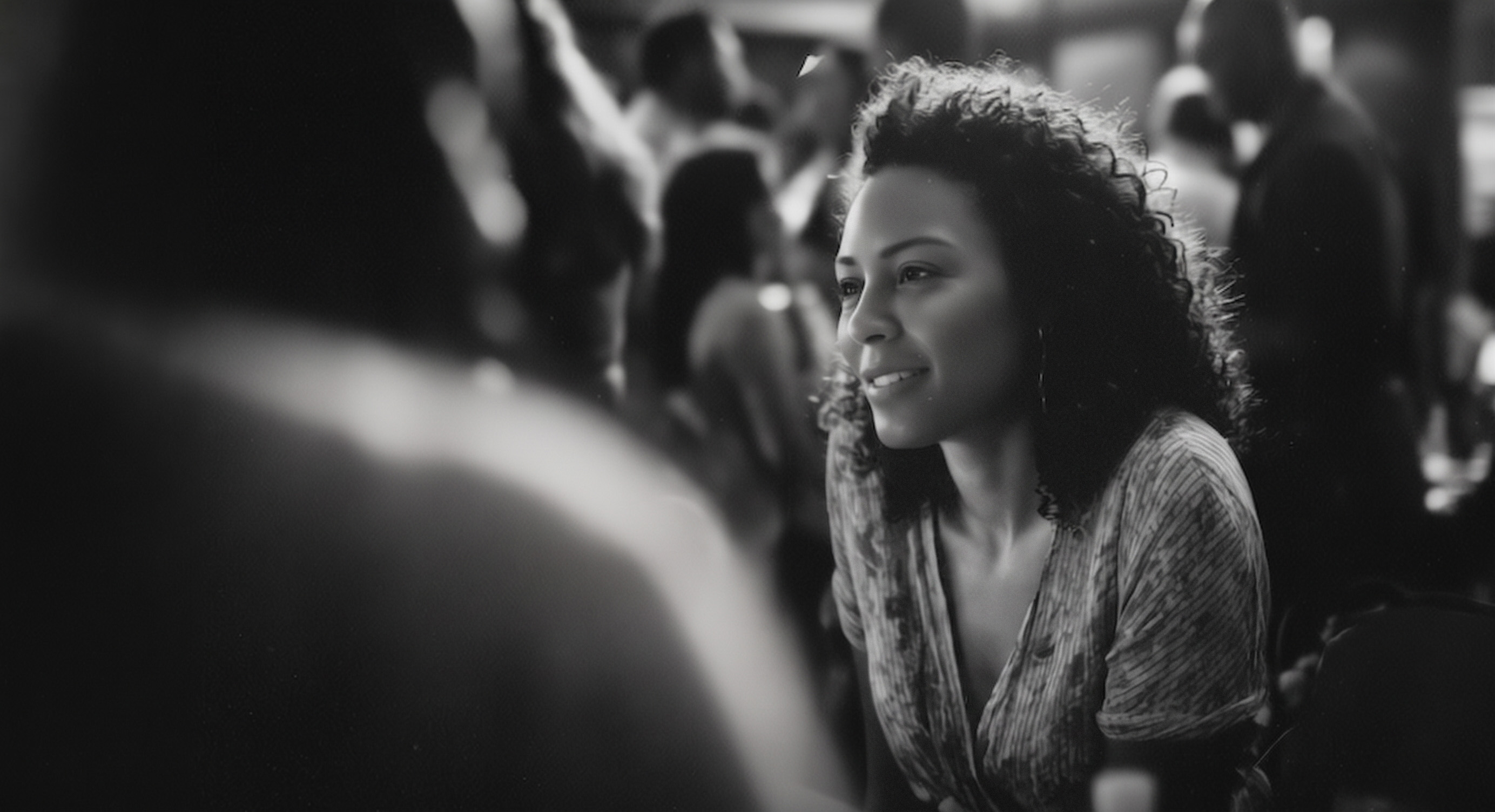 Black-and-white photograph: a woman with curly hair, smiling, leans in to listen to someone across from her in a crowded room