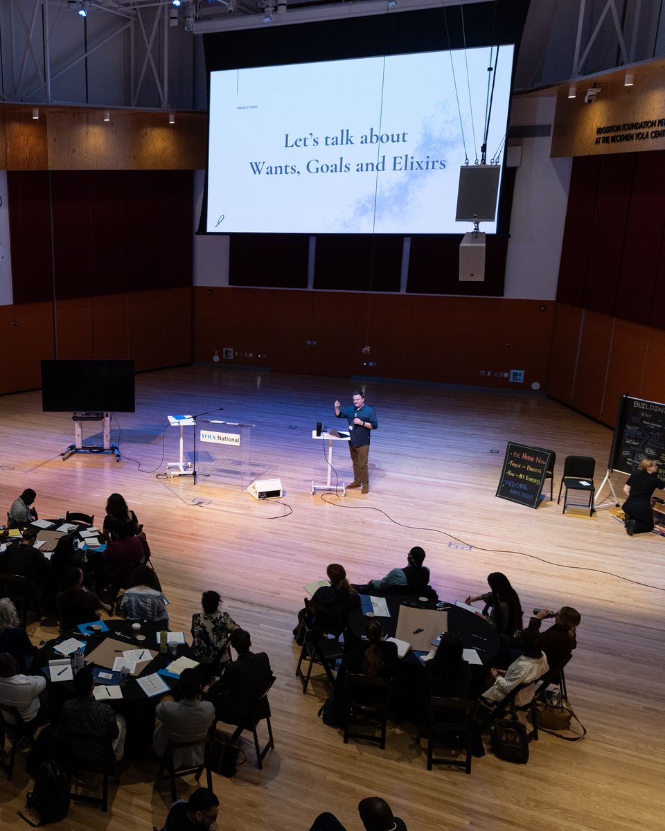A large hall of people at round tables during a workshop, a presenter on stage beneath a slide reading “Let’s talk about Wants, Goals and Elixirs.”