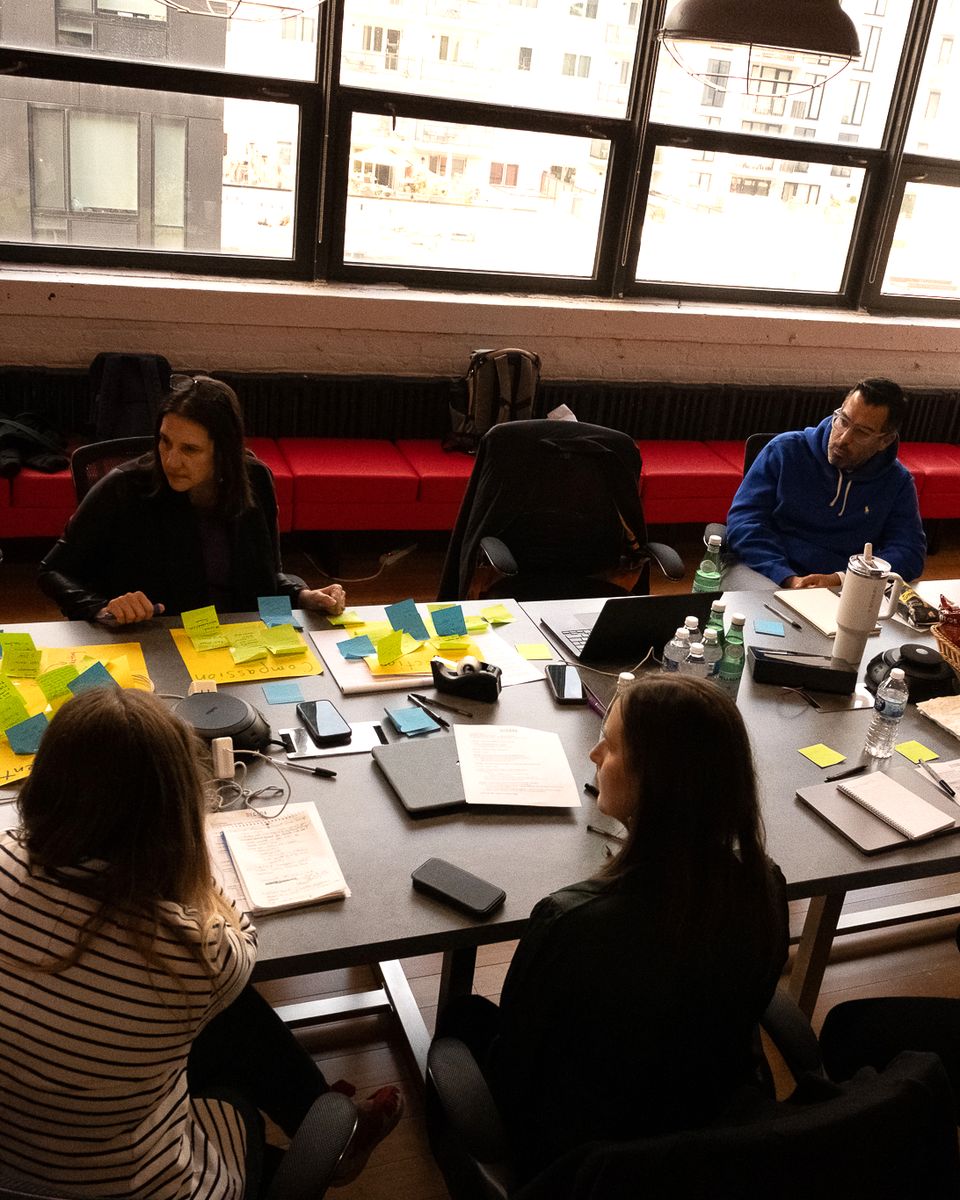 Six people work around a long table covered in sticky notes, laptops and papers, in a room with tall windows.