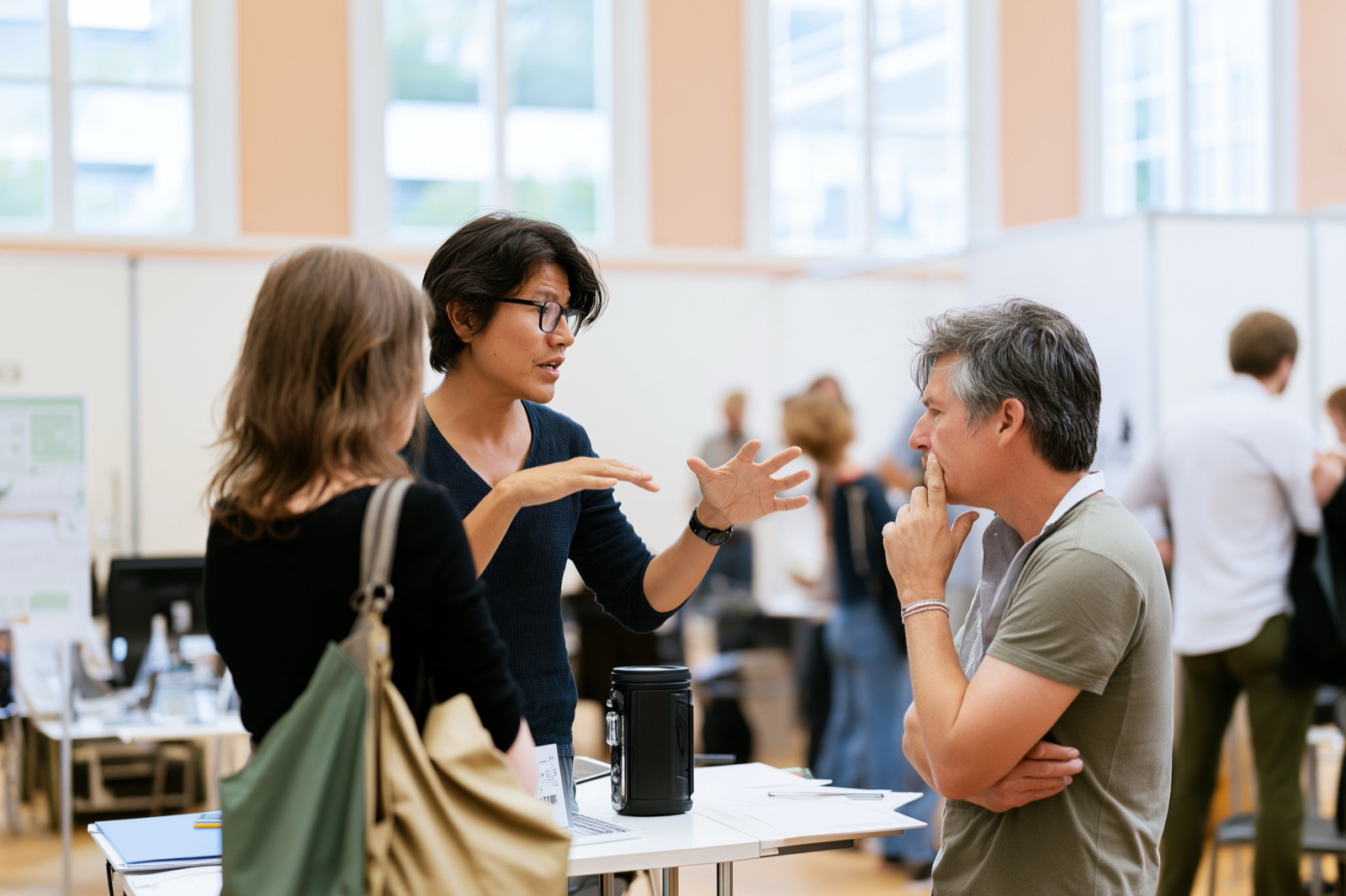 A founder mid-explanation at a trade-fair booth, both hands shaping the idea in the air, while a visitor listens with a hand at his chin, not yet convinced.
